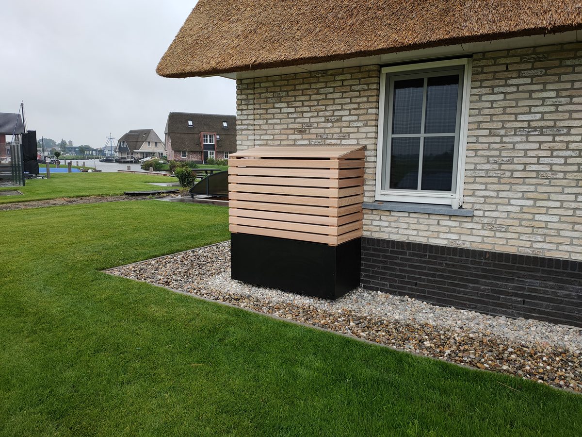 Houten ombouw op maat voor een warmtepomp, naast een woning met rieten dak